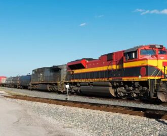 Ein langer Güterzug mit rot-gelber Diesellokomotive auf den Gleisen unter blauem Himmel.