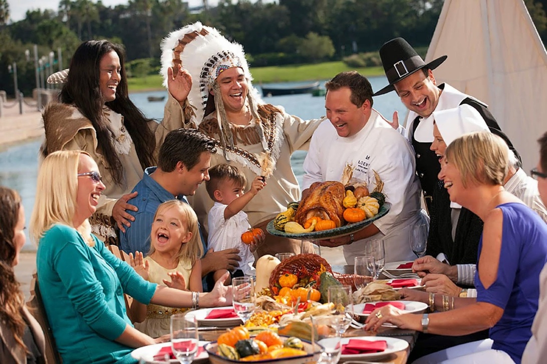 Eine festliche Thanksgiving-Versammlung mit Menschen, die um einen reich gedeckten Tisch mit traditionellen Gerichten sitzen. Ein gebratener Truthahn ist prominent zu sehen, während die Teilnehmer, einige in Kostümen der amerikanischen Ureinwohner und Pilger, lächeln und gemeinsam feiern. Die Szene spielt sich im Freien in der Nähe von Wasser ab und schafft eine fröhliche Feiertagsatmosphäre, die an die historische Thanksgiving-Erzählung erinnert.